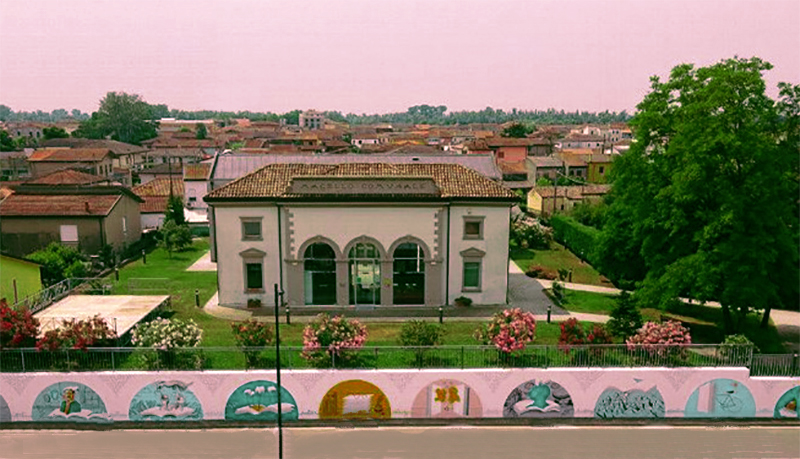 Veduta aerea di un edificio storico a Porto Viro, circondato da giardini e murales colorati lungo la strada.